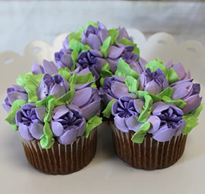 A CC-091 Gold or Chocolate cupcake with white filling is displayed on a white scalloped-edge plate, decorated with colorful buttercream flowers—purple petals—and green leaves.