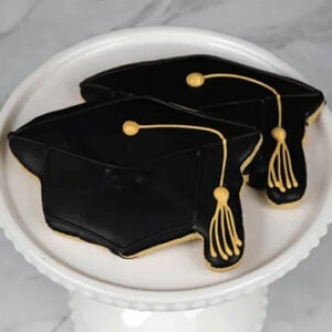 Two black frosted cookies with a gold tassel (Product: GC-001) are displayed on a white decorative cake stand against a marble background.