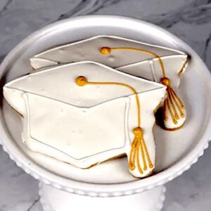 Two white frosted cookies with a gold tassel (Product: GC-002) are displayed on a white decorative cake stand against a marble background.