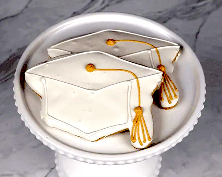Two white frosted cookies with a gold tassel (Product: GC-002) are displayed on a white decorative cake stand against a marble background.