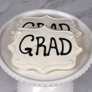 Two white frosted cookies with "GRAD" in black icing (Product: GC-004) are displayed on a white decorative cake stand against a marble background.