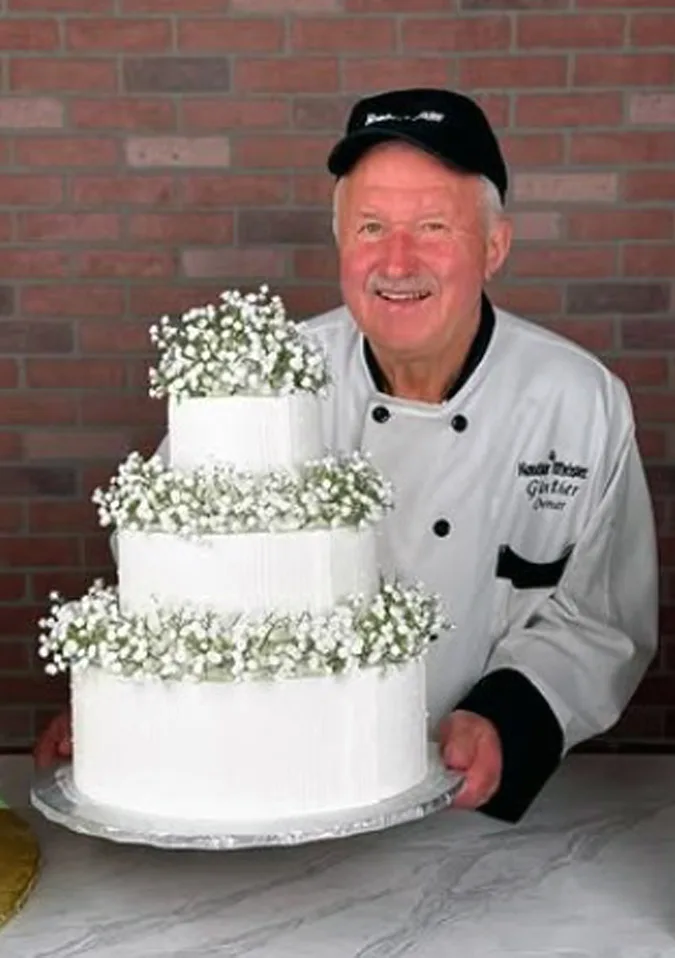A smiling man in a chef’s uniform and black cap holds a three-tiered white cake decorated with small white flowers, standing in front of a brick wall that gives the scene a warm, home-inspired feel.