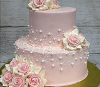 A two-tiered pink cake decorated with white and pink buttercream roses and small white pearl-like icing accents, perfect for a home celebration, displayed on a golden board against a wooden background.