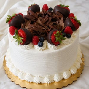 The BD-018D Triple Berry Cake features a white frosted finish, chocolate shavings, chocolate-covered strawberries, raspberries, and blueberries on a scalloped gold cake board.