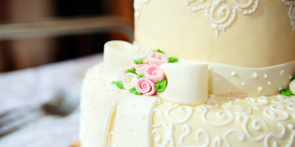 Close-up of an elegant white cake, perfect for a home celebration, adorned with intricate icing designs, white ribbons, and pastel pink and white sugar roses with green leaves.