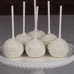 Six DB-000B Gold Cake Pops, each on a stick with gold coating and sprinkles, are arranged on a white beaded-edge cake stand, set on a marble surface against a dark brick background.