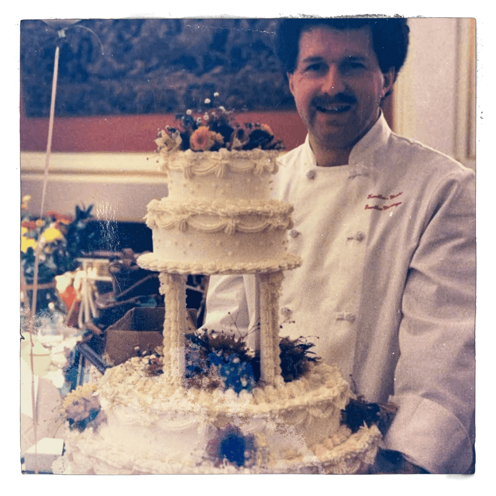 A smiling man in a white chef’s coat stands behind a large, ornate three-tiered cake decorated with white icing and colorful flowers in a bright indoor setting.