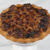 Pie-Pecan sits on a round wooden surface, topped with chocolate cream swirls, drizzled with chocolate sauce, and sprinkled with nuts. The background is blurred with red and festive decorations.