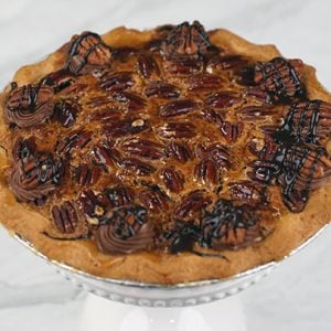 Pie-Pecan sits on a round wooden surface, topped with chocolate cream swirls, drizzled with chocolate sauce, and sprinkled with nuts. The background is blurred with red and festive decorations.