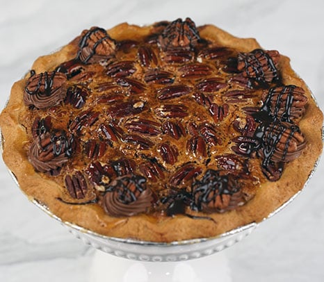 Pie-Pecan sits on a round wooden surface, topped with chocolate cream swirls, drizzled with chocolate sauce, and sprinkled with nuts. The background is blurred with red and festive decorations.