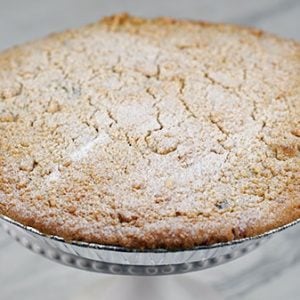 A Pie-Apple with a golden, crumbly topping rests in a foil pan on a tabletop, set against a blurred festive background with red and holiday decorations.