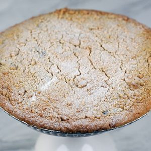 A Pie-Blueberry with a golden brown crumb topping rests in a foil pie tin on a round surface, with blurred festive decorations in the background.