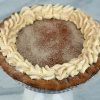 A Pie-Pumpkin with a golden-brown crust is finished with whipped cream swirls and a sprinkle of cinnamon or nutmeg on top, presented on a dark surface with a red cloth in the background.