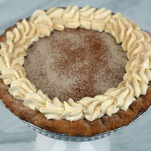 A Pie-Pumpkin with a golden-brown crust is finished with whipped cream swirls and a sprinkle of cinnamon or nutmeg on top, presented on a dark surface with a red cloth in the background.
