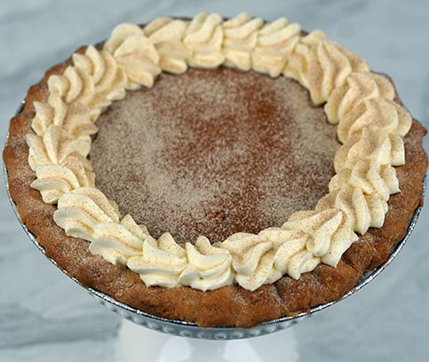 A Pie-Pumpkin with a golden-brown crust is finished with whipped cream swirls and a sprinkle of cinnamon or nutmeg on top, presented on a dark surface with a red cloth in the background.