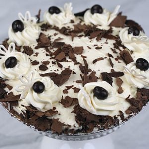 The Pie-Chocolate Cream is a round chocolate pie topped with whipped cream swirls, chocolate shavings, and dark chocolate pieces, displayed on a glass cake stand against a white background.