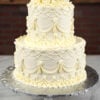 WD-104 is a two-tier white wedding cake featuring intricate vintage-style piping and elegant draped details. White buttercream roses crown the top, creating a classic and sophisticated look.