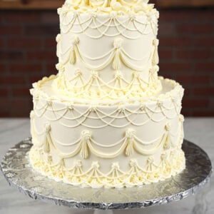 WD-104 is a two-tier white wedding cake featuring intricate vintage-style piping and elegant draped details. White buttercream roses crown the top, creating a classic and sophisticated look.