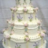 WD-107 is a four-tier ivory wedding cake decorated with soft pink buttercream roses, delicate lavender flowers, green leaves, and classic draped piping. The intricate floral details give the cake a romantic, vintage-inspired look.