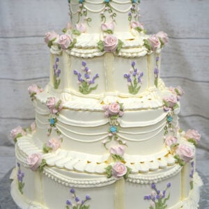WD-107 is a four-tier ivory wedding cake decorated with soft pink buttercream roses, delicate lavender flowers, green leaves, and classic draped piping. The intricate floral details give the cake a romantic, vintage-inspired look.