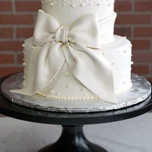 WD-108 is a two-tier white wedding cake decorated with pearl accents and an oversized white bow wrapped around the tiers.