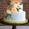WD-109 is a three-tier wedding cake with soft ivory and pale blue icing, gold accents, and a cascading arrangement of blush, peach, and white roses with greenery.