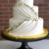 WD-110 is a three-tier white wedding cake featuring elegant draped icing, delicate pearl accents, and cascading white flowers with greenery.