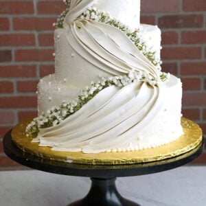 WD-110 is a three-tier white wedding cake featuring elegant draped icing, delicate pearl accents, and cascading white flowers with greenery.
