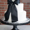 WD-113 is a three-tier white wedding cake with subtle pearl accents and a dramatic oversized black bow cascading down the front.
